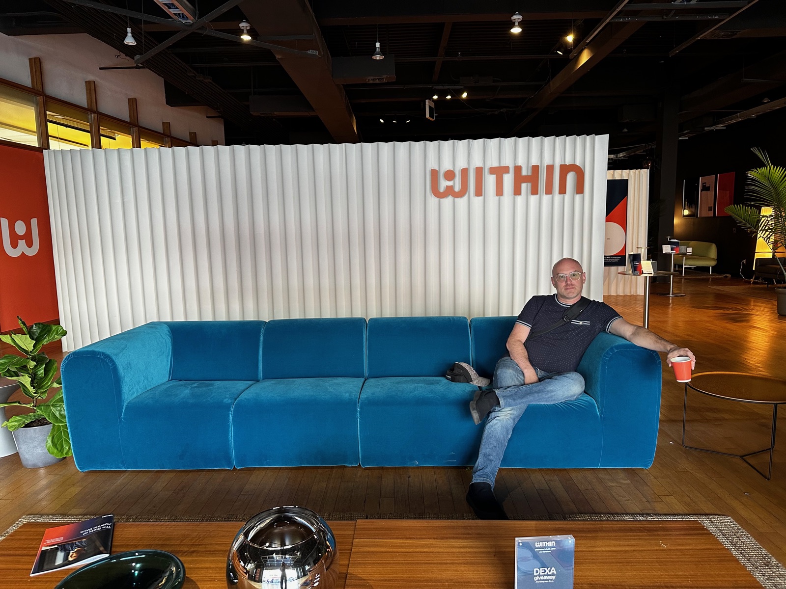 The author sits with a coffee on a long blue velvet couch in front of a fluted white wall reading WITHIN, in a Palo Alto pop-up that looks like a furniture showroom