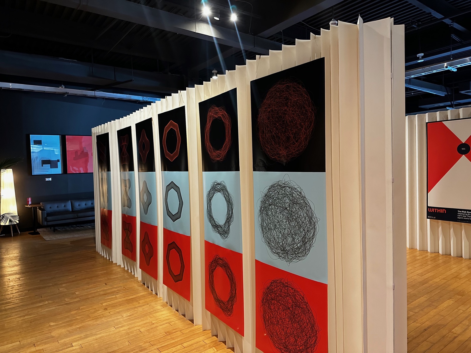 A long fluted wall of WITHIN posters receding into the room, each a red, black, and cream panel of tangled scribble-ball shapes that read as art more than data