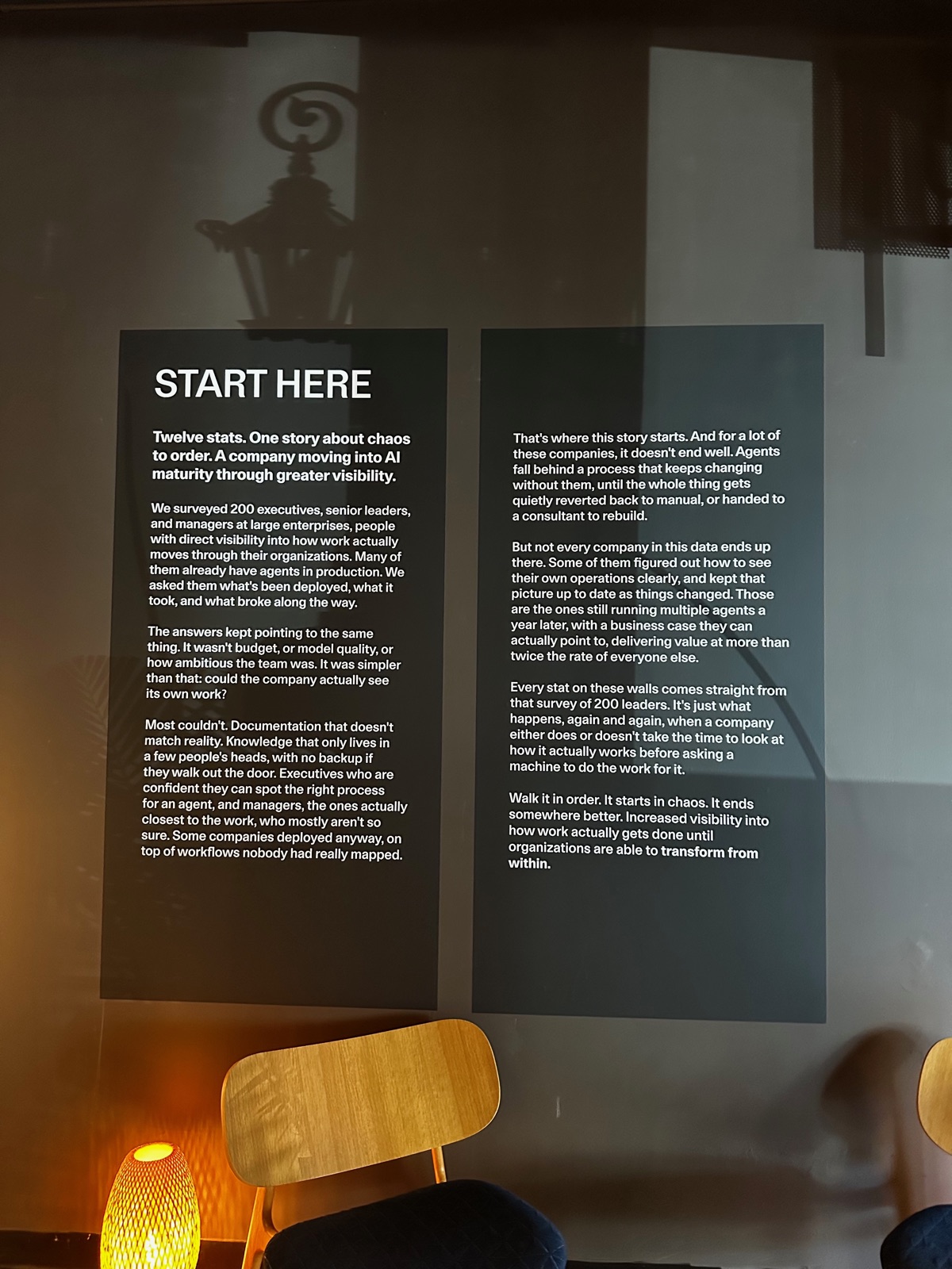 A backlit wall of small-print exhibition text headed START HERE, opening 'Twelve stats. One story about chaos to order,' in the unmistakable cadence of AI-written copy